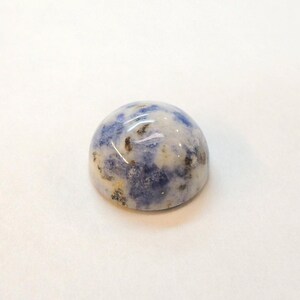 Op de afbeelding: Een gepolijste, cabochon-geslepen edelsteen met een afgeronde koepelvorm. De steen heeft een mix van blauwe, witte en bruine kleuren, waardoor een gespikkeld patroon ontstaat. De edelsteen is geplaatst tegen een effen witte achtergrond.