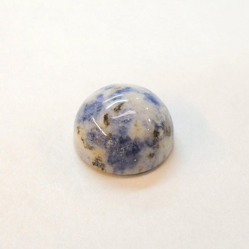 Op de afbeelding: Een gepolijste, cabochon-geslepen edelsteen met een afgeronde koepelvorm. De steen heeft een mix van blauwe, witte en bruine kleuren, waardoor een gespikkeld patroon ontstaat. De edelsteen is geplaatst tegen een effen witte achtergrond.