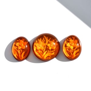 May include: Three amber-colored glass cabochons with a floral design. The cabochons are oval and round in shape. The flowers are carved into the glass and are a light orange color.