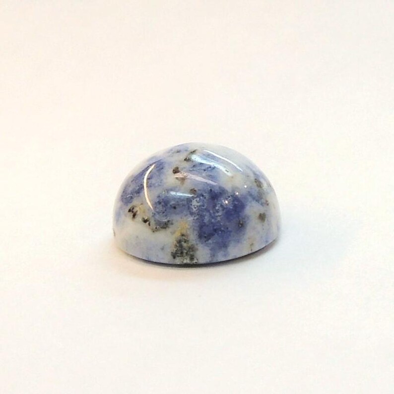 Op de afbeelding: Een gepolijste, cabochon-geslepen edelsteen met een afgeronde koepelvorm. De steen heeft een mix van blauwe, witte en grijze kleuren, met kleine donkere vlekjes. Het oppervlak is glad en glanzend en reflecteert licht.