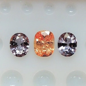 May include: Three faceted gemstones, one orange, one purple and one pink, are displayed on a white background.