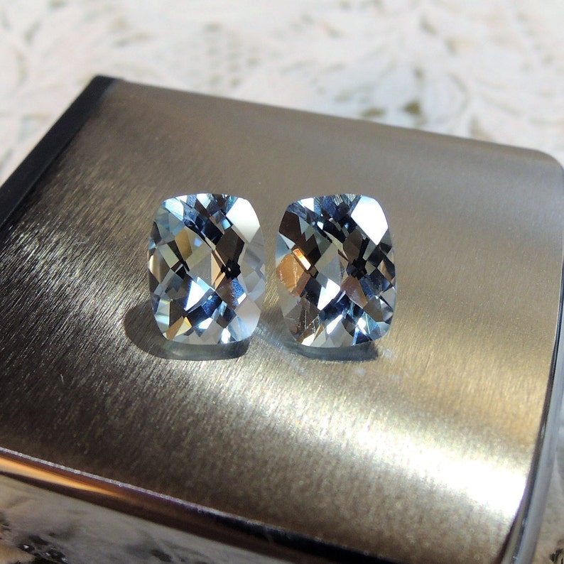 Puede incluir: Dos piedras preciosas de aguamarina de corte coj&iacute;n de color azul claro sobre un fondo plateado.