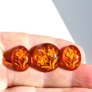 May include: Three amber-colored glass buttons with a floral design. The buttons are oval-shaped and have a raised design of flowers and leaves.