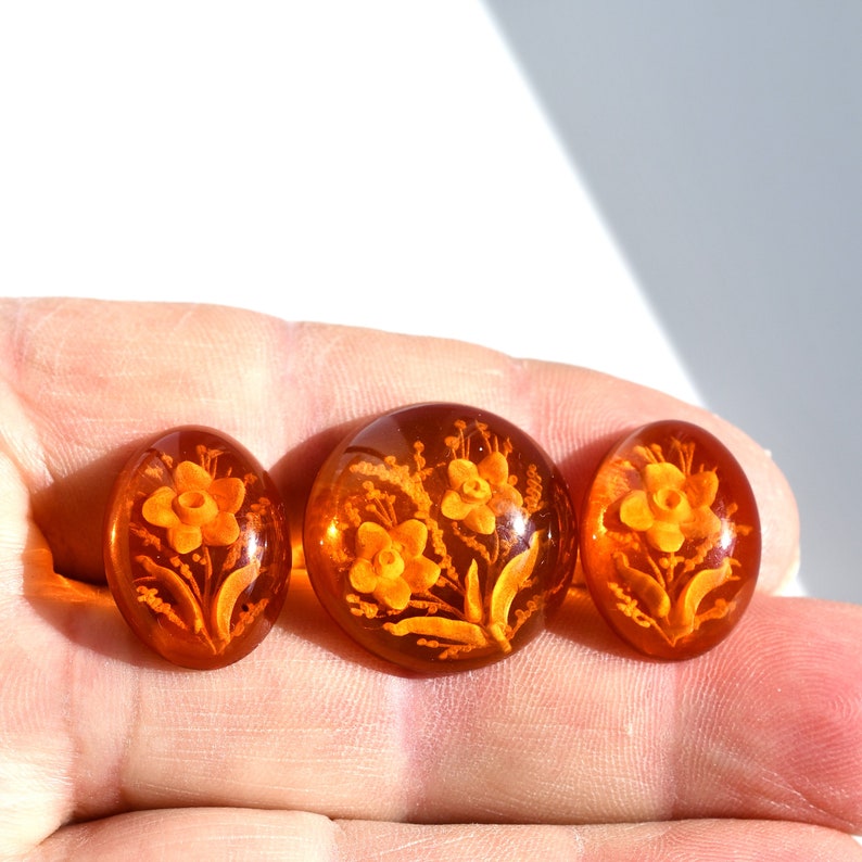 May include: Three amber-colored glass buttons with a floral design. The buttons are oval-shaped and have a raised design of flowers and leaves.