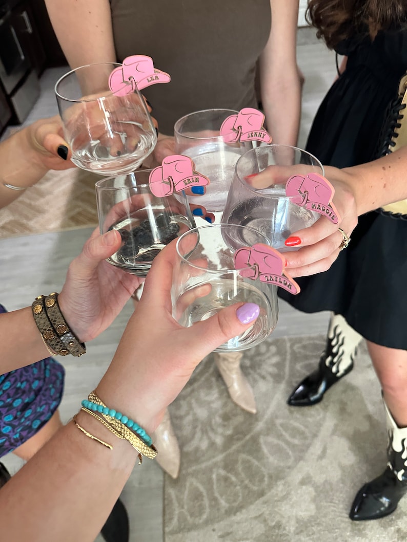 May include: Five clear glass cups with pink cowboy hat shaped drink markers. The markers have the names "LEA", "JENNY", "ERIN", "MADDOX", and "TAYLOR" written on them.