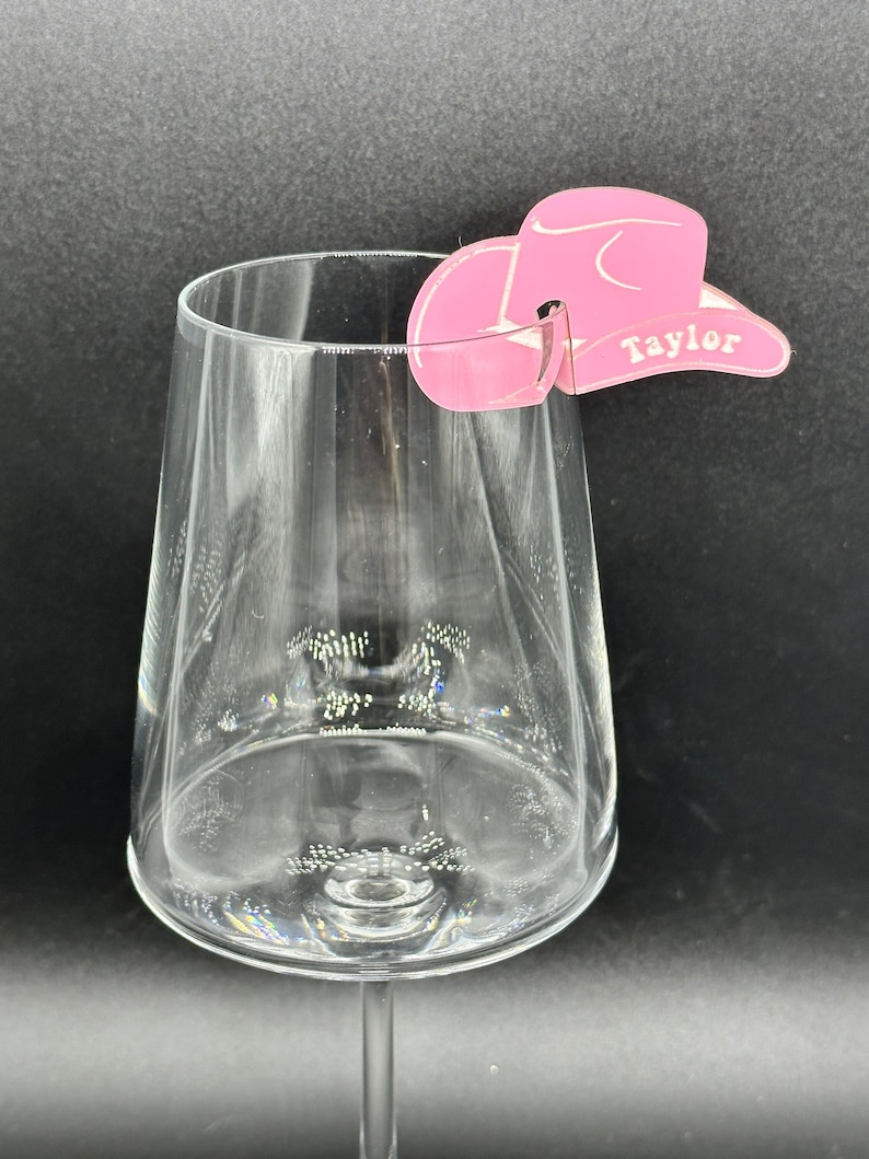 May include: Pink cowboy hat shaped wine glass charm with the name "Taylor" written on the brim.