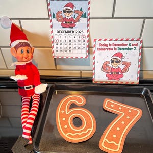 May include: A Christmas elf doll in a red suit and striped legs stands next to a baking sheet with gingerbread numbers 6 and 7. Two signs read "December 2025" and "Today is December 6 and tomorrow is December 7."