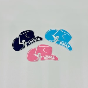 May include: Three foam cowboy hats with names written on them. The hats are blue, pink, and black. The names are Caitlyn, Kelly, and Becca.