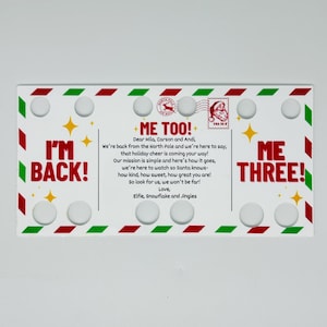 Pode incluir: Um letreiro retangular branco com bordas vermelhas e verdes, com as frases "I'M BACK!", "ME TOO!" e "ME THREE!". O letreiro inclui texto sobre a alegria das festas e é decorado com estrelas e ímãs circulares.