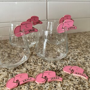 May include: Pink cowboy hat shaped drink markers with names written on them. The names are Erin, Lea, Bailie, Maggie, Emily, and Jenny.