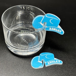 May include: Two blue cowboy hat shaped drink markers with the name "Ashley" written on them. The markers are made of a hard plastic material and are designed to be placed on the rim of a glass.