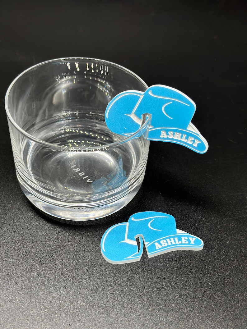 May include: Two blue cowboy hat shaped drink markers with the name "Ashley" written on them. The markers are made of a hard plastic material and are designed to be placed on the rim of a glass.