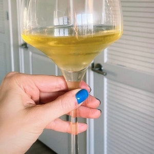 May include: A clear glass wine glass filled with a light yellow liquid. A pink cowboy hat with the name "Jenny" written on it is attached to the rim of the glass.