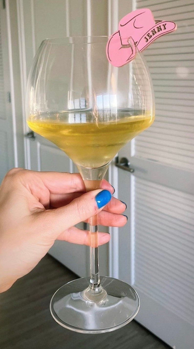 May include: A clear glass wine glass filled with a light yellow liquid. A pink cowboy hat with the name "Jenny" written on it is attached to the rim of the glass.