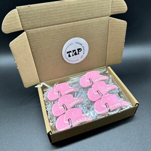 May include: A box filled with pink cowboy hats with names written on them. The box is open and the hats are visible. The box has a sticker on it that says "Laser Design Co. TAP".