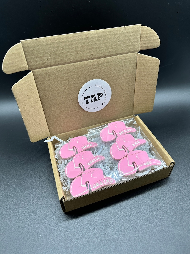 May include: A box filled with pink cowboy hats with names written on them. The box is open and the hats are visible. The box has a sticker on it that says "Laser Design Co. TAP".