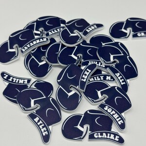 May include: A pile of navy blue felt hats with white lettering. The hats are shaped like graduation caps and have different names written on them, including Savannah, Ally, Abby, Emily, Lexi, Millie, Sophie, Claire, and Riley.