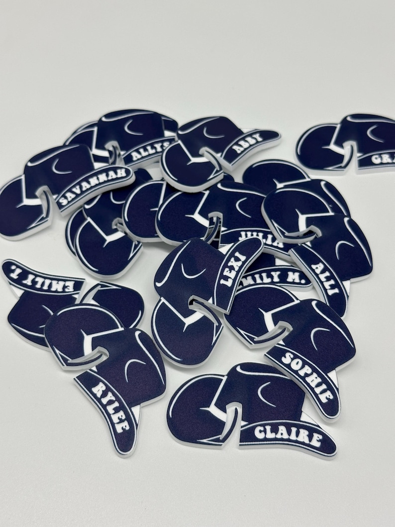 May include: A pile of navy blue felt hats with white lettering. The hats are shaped like graduation caps and have different names written on them, including Savannah, Ally, Abby, Emily, Lexi, Millie, Sophie, Claire, and Riley.
