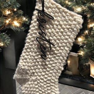 May include: A white knitted Christmas stocking with a black metal tag that says "Kristen" hanging from it.