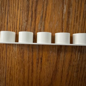 May include: A white shelf with five cylindrical objects. The objects are evenly spaced along the shelf. The shelf and cylinders are white and made of a smooth material. The background is a wooden surface with a visible grain.