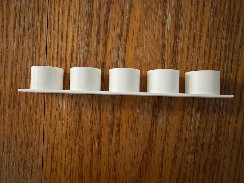 May include: A white shelf with five cylindrical objects. The objects are evenly spaced along the shelf. The shelf and cylinders are white and made of a smooth material. The background is a wooden surface with a visible grain.