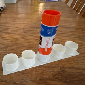 May include: A white 3D-printed glue stick holder with four circular compartments and a central space for a glue stick. The glue stick is orange, blue, and white. The holder sits on a wooden table.