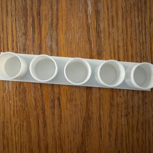 May include: A white, 3D-printed spice rack with six circular openings. The rack is rectangular and designed to hold small containers. The object is made of a light-coloured plastic and sits on a wooden surface.