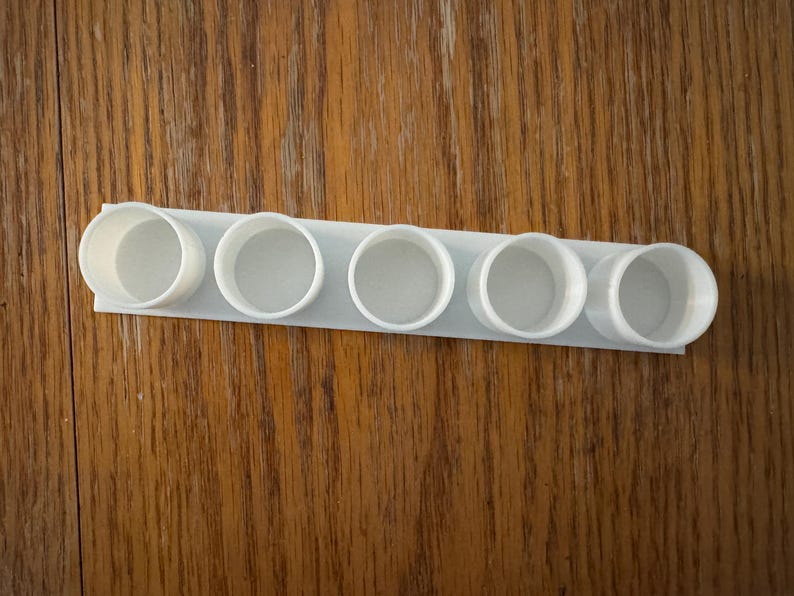 May include: A white, 3D-printed spice rack with six circular openings. The rack is rectangular and designed to hold small containers. The object is made of a light-coloured plastic and sits on a wooden surface.
