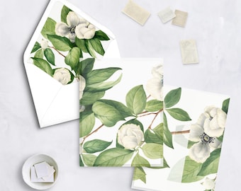 May include: White stationery set featuring a floral design with white flowers and green leaves. The set includes an envelope, two cards, and stamps. The floral design is printed on the envelope and cards.