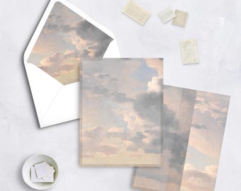 May include: Stationery set featuring a sky-themed design. Includes a white envelope with a sky-themed interior, several notecards, and stamps. The notecards and envelope interior display a soft, pastel-colored cloudscape. A small white bowl holds additional stamps.