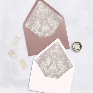 May include: Two open envelopes, one dusty rose and one white, with ornate floral patterned linings. Several vintage postage stamps and a small dish with stamps are also visible, suggesting stationery or wedding invitations.
