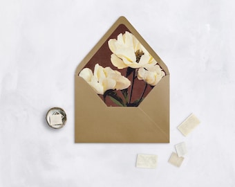May include: A gold-colored envelope with a floral design inside, featuring white flowers against a dark brown background. A small container with tiny white squares and several small rectangular stickers are also present.