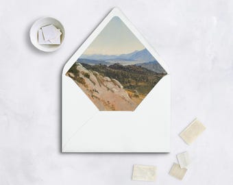 May include: White envelope with a landscape painting visible inside. The painting depicts a mountain range and valley in shades of green, brown, and blue. Several stamps and a small bowl of stamps are scattered around the envelope on a white surface.