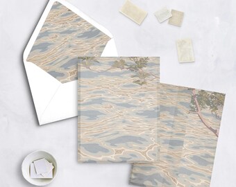 May include: Stationery set featuring a watercolor design of water with light blue and tan hues. The set includes an envelope, notecards, and stamps. The envelope's interior and the notecards have the same water design. A small white bowl holds additional stamps.