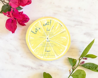 May include: A yellow lemon slice with a blue and white menu on it. The menu says "Let's eat!" and lists the food items: Caesar Salad, Grilled Prawns, Parmesan Crostini, Veal Medallions, Wild Mushroom Risotto, Herbed Broccoli, Tiramisu, Dark Chocolate Gelato.
