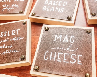 May include: A set of six wooden signs with brown leatherette tops. Each sign has white text on the leatherette. The signs read: "Grilled White Fish Thai BBQ Glaze", "BBQ Baked Beans", "Dessert Fruit Cobbler Cookies Station", "Mac and Cheese", "PUD", and "S".