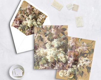 May include: Stationery set featuring floral artwork. The set includes a white envelope with a floral interior, two cards with a similar design, and several small, rectangular stamps. The artwork showcases white and purple flowers with green leaves against a tan background.