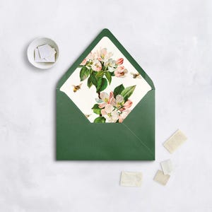 May include: A dark green envelope with a floral design on the inside, featuring pink and white blossoms, green leaves, and bees. Several stamps and a small white bowl with stamps are also present.