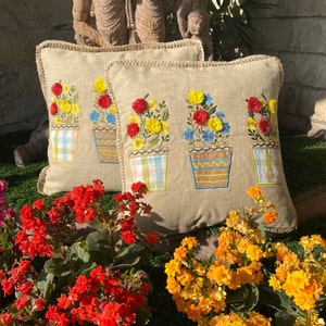 May include: Three embroidered throw pillows with a beige linen fabric. Each pillow features a design of three potted flowers with red, yellow, and blue flowers. The pillows have a crocheted trim.