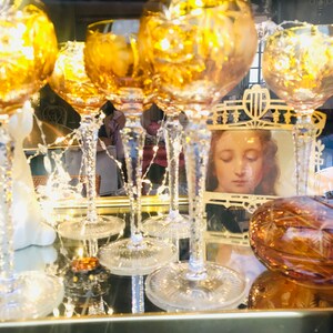 Puede incluir: Una colecci&oacute;n de seis copas de vidrio &aacute;mbar con intrincados dise&ntilde;os tallados, expuestas sobre una superficie de espejo con un marco dorado. Las copas est&aacute;n iluminadas desde el interior, creando un brillo c&aacute;lido. Un peque&ntilde;o cuadro enmarcado de una mujer con los ojos cerrados es visible en el fondo.