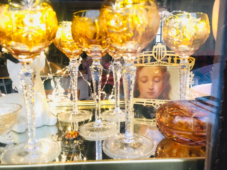 Puede incluir: Una colecci&oacute;n de seis copas de vidrio &aacute;mbar con intrincados dise&ntilde;os tallados, expuestas sobre una superficie de espejo con un marco dorado. Las copas est&aacute;n iluminadas desde el interior, creando un brillo c&aacute;lido. Un peque&ntilde;o cuadro enmarcado de una mujer con los ojos cerrados es visible en el fondo.