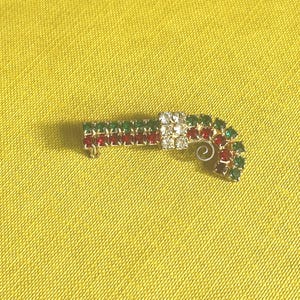 Könnte beinhalten: Eine Brosche in Form einer Zuckerstange mit abwechselnden Reihen aus roten und grünen Strasssteinen, akzentuiert durch ein mittleres Band aus klaren weißen Strasssteinen und ein kleines Metallspiraldetail am gebogenen Ende, in Goldton.