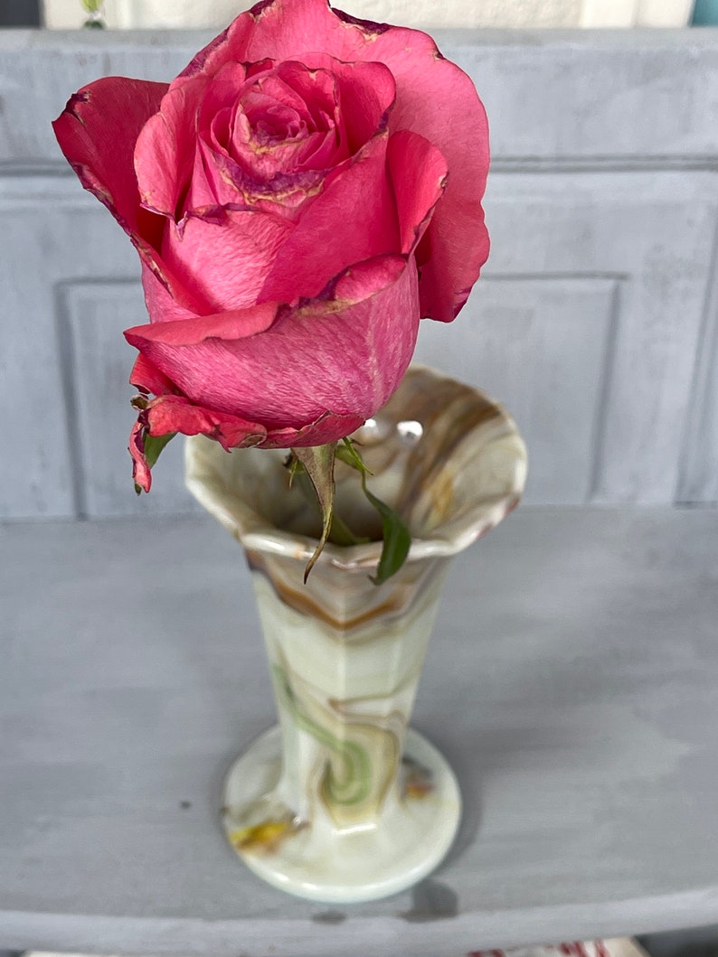 May include: A single pink rose with a green stem in a swirl patterned glass vase with brown, green, and white colors.