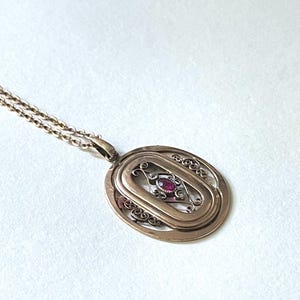 Könnte beinhalten: Eine goldene Halskette mit einem Anhänger im Filigran-Design und einem roten Edelstein in der Mitte. Der Anhänger ist oval geformt und hängt an einer zarten Kette.