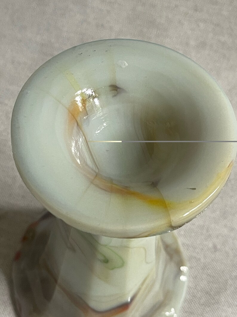 May include: A white glass vase with a swirl pattern in shades of brown, orange, and green. The vase has a wide, round opening and a narrow base.
