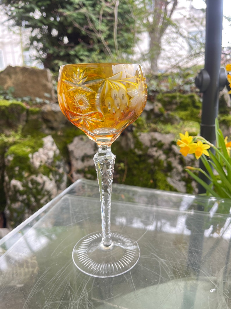 Puede incluir: Un vaso alto de color &aacute;mbar con intrincados dise&ntilde;os de vidrio cortado. El vaso tiene un tallo largo y delgado y un cuenco ancho y redondo. El vaso est&aacute; sobre una superficie de vidrio transparente.