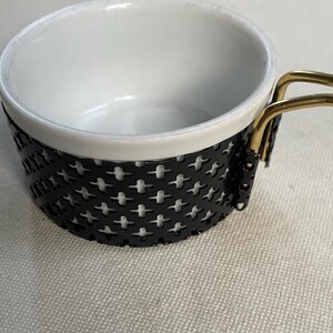 Könnte beinhalten: Eine weiße Keramiktasse mit einem schwarzen Metallgriff und einem schwarzen Metallgitterdesign um den Boden. Der Griff hat goldene Akzente.