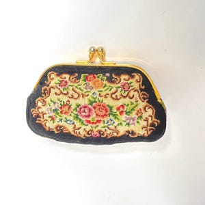 Könnte beinhalten: Eine schwarze Vintage-Geldbörse mit floralem Nadeldesign. Die Geldbörse hat einen goldfarbenen Verschluss mit Perlenakzenten. Das Nadeldesign zeigt rosa, rote und orangefarbene Rosen mit grünem und blauem Laub.