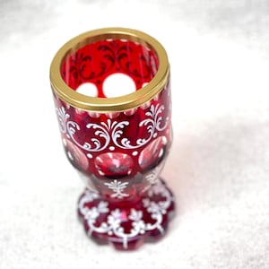 Könnte beinhalten: Ein tiefroter Glaskelch mit goldfarbenem Rand und filigranen, weiß bemalten Mustern. Das Gefäß hat ein facettiertes, kreisförmiges Design mit klaren Glasausschnitten und einen gewellten Fuß, der mit passenden weißen Blumenmotiven verziert ist.