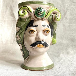 Könnte beinhalten: Eine Keramikvase in Form eines menschlichen Kopfes mit grün-weißen Maldetails. Die Vase hat einen Schnurrbart und eine grün-schwarz gemusterte Krone.
